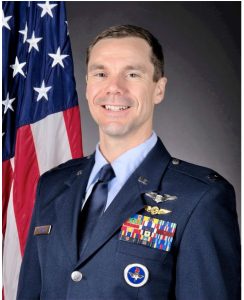 A young man in a blue military uniform and dark hair stands smiling against a dark gradient background with an American flag on the left. 