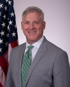 A grey-haired man in a grey suit and light blue tie stands smiling. 